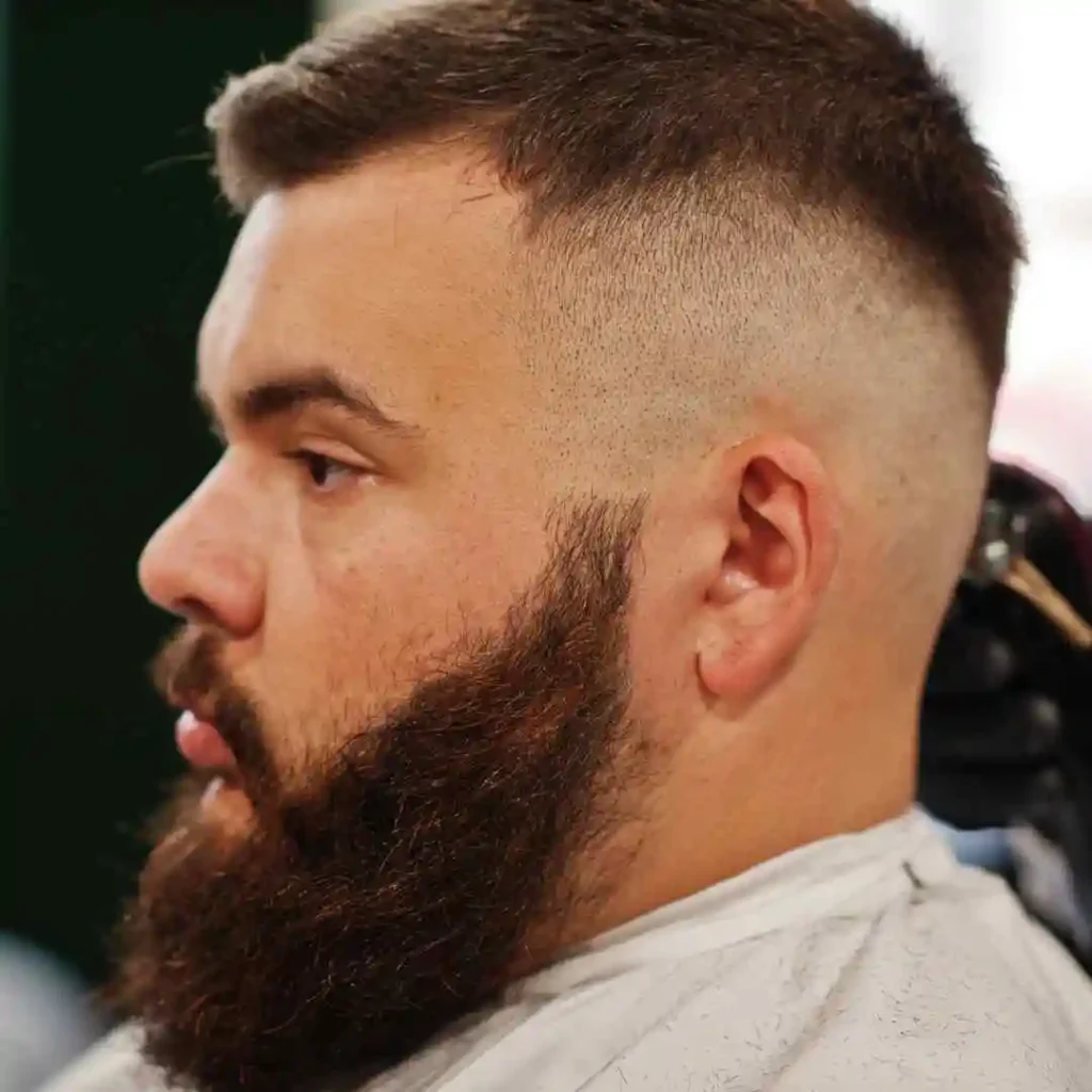 A man with a thick beard and faded haircut sits in a barber's chair, looking to the side with a focused expression.