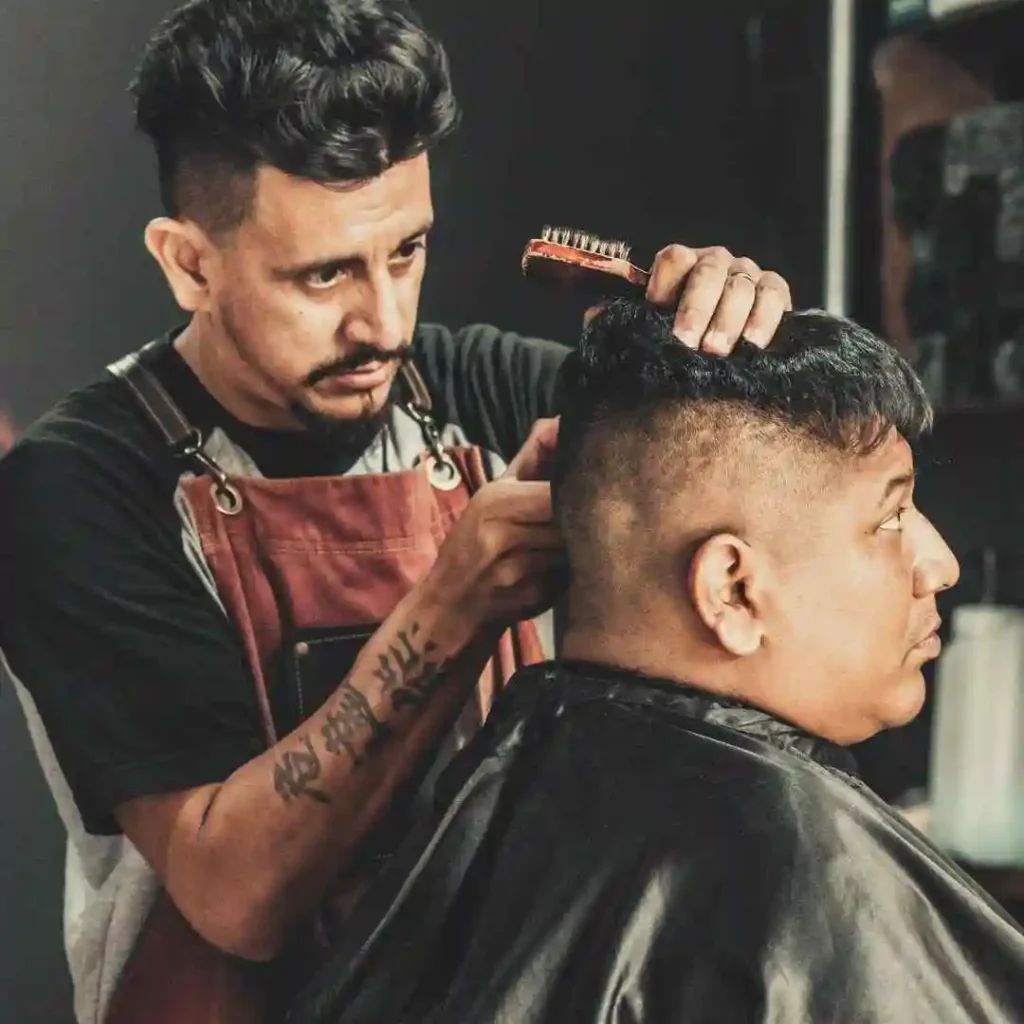 A barber in an apron carefully styles the hair of a client draped in a black cape, focused in a modern barbershop setting.