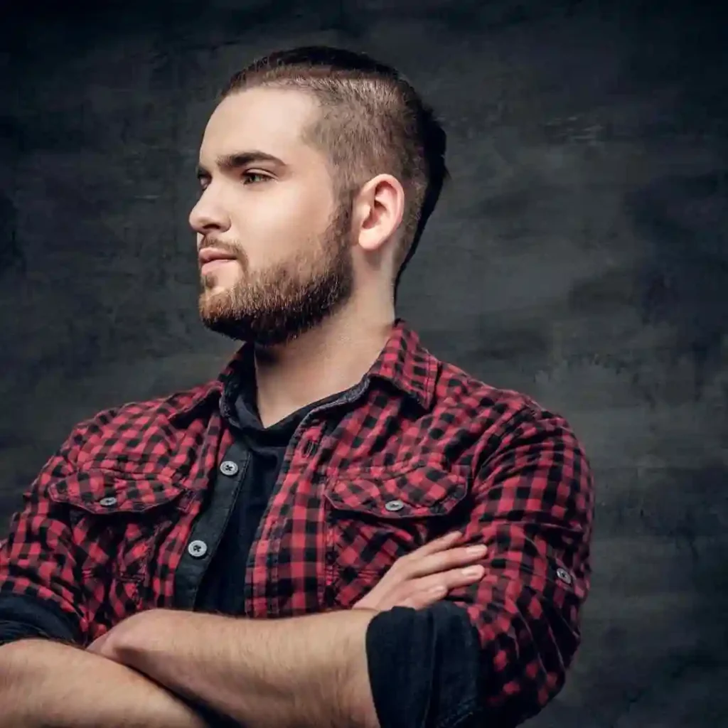 A man with crossed arms stands against a dark, textured background, wearing a red and black checkered shirt over a black tee, showcasing a stylish MEN'S CROP & CREW CUTS hairstyle.