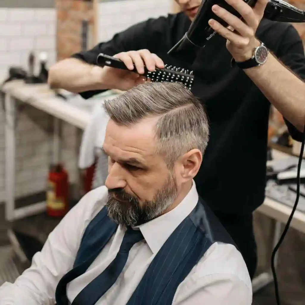 A man with a beard sits in a barbershop chair, as a stylist brushes his hair with a comb and blow dryer in use.