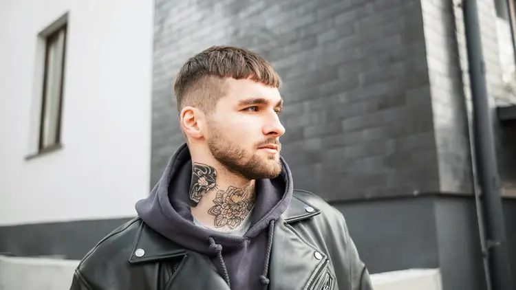 A young man in a black leather jacket and hoodie stands outdoors against a modern, textured gray wall. He has visible neck tattoos, showcasing a trendy men hairstyle undercut.