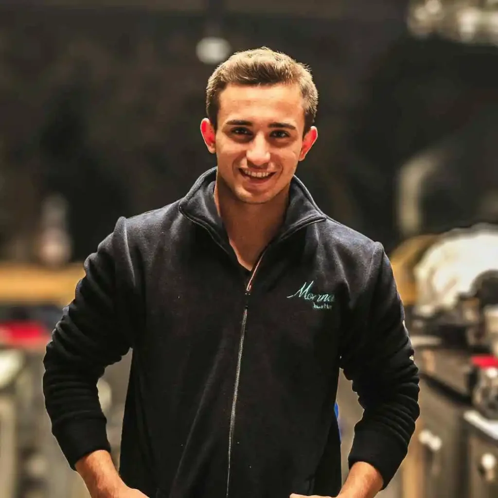 A young man smiles confidently, wearing a dark zip-up jacket, with kitchen equipment visible in the background.