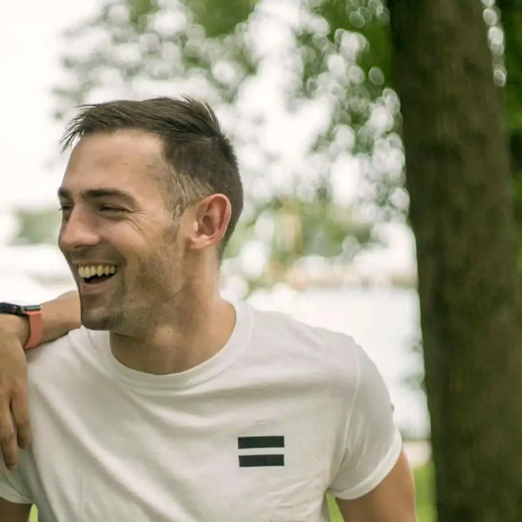 A person wearing a white t-shirt with a black striped design stands outdoors, with trees in the background and a blurred lake visible, showcasing MEN HAIRSTYLES UNDERCUT.
