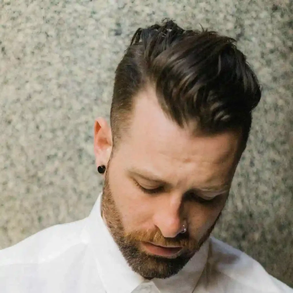 A person with dark, slicked-back hair, showcasing a MEN HAIRSTYLES UNDERCUT, and visible earrings, wearing a white shirt, stands against a textured stone background.