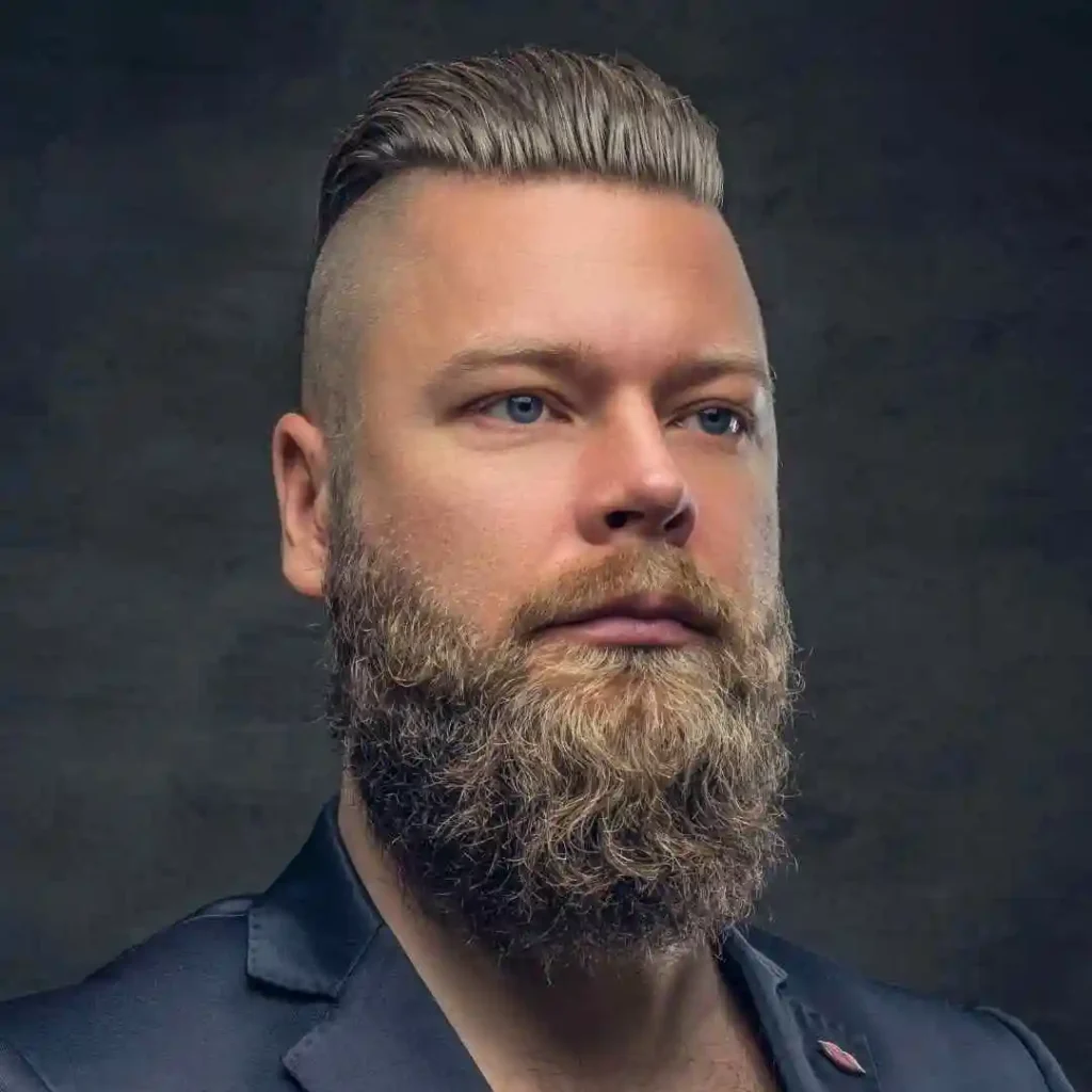 A close-up portrait of a man with a well-groomed beard and a stylish MEN'S CROP & CREW CUTS hairstyle, wearing a dark suit against a moody background.