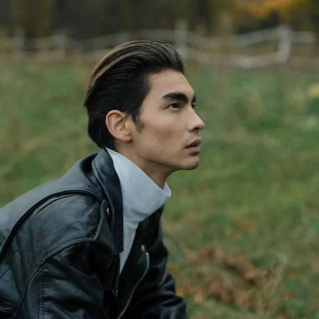 20 Trendy Options for Men Hairstyles Undercut 3 A person with MEN HAIRSTYLES UNDERCUT, wearing a black leather jacket and white shirt, sits in a grassy field surrounded by autumn foliage and a wooden fence in the background.