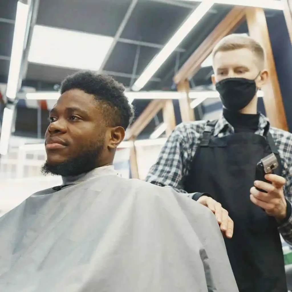 A barber works on a client in a stylish barbershop, both wearing masks. The client is draped with a barber cape.