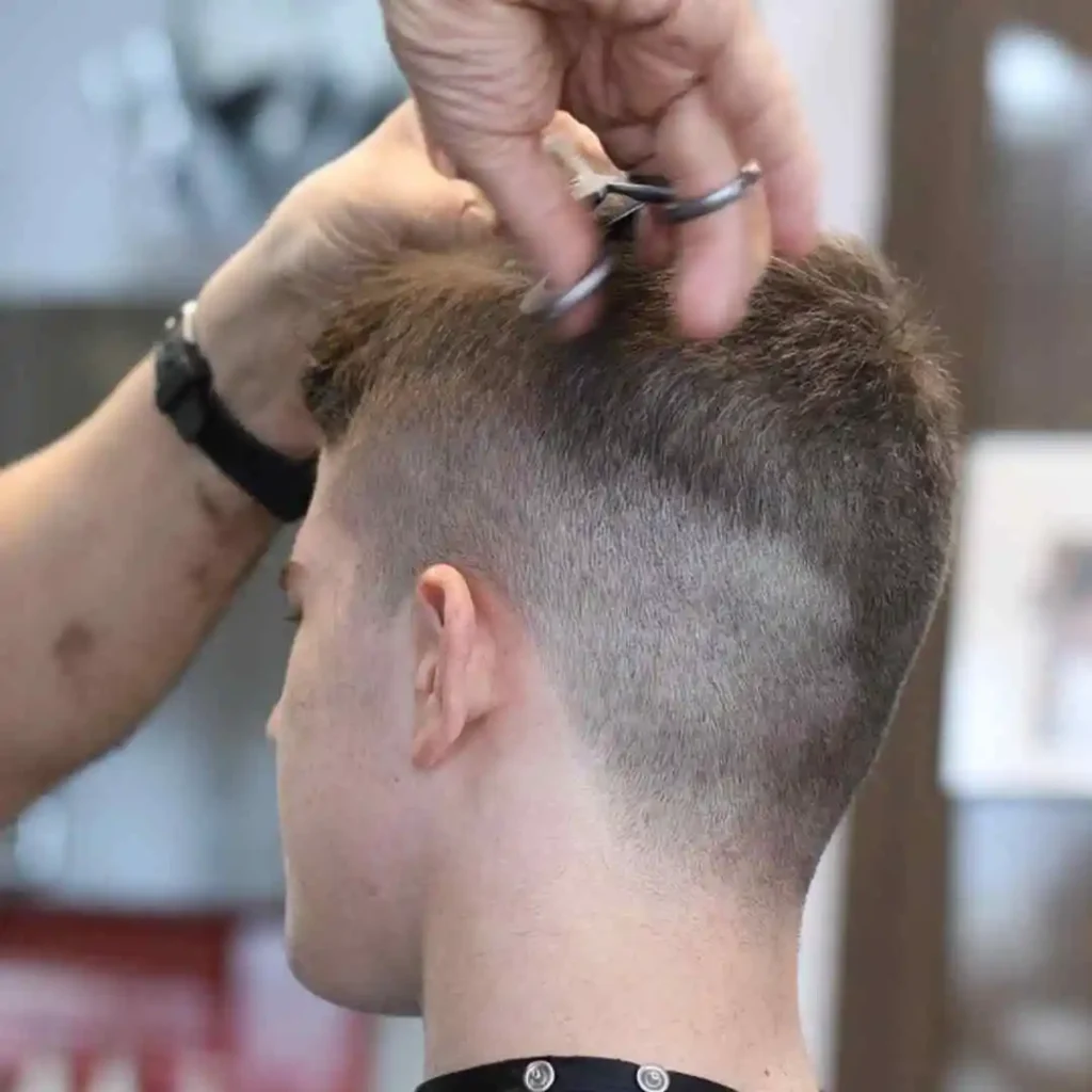 A close-up of a barbershop scene, showing a stylist trimming the back of a young man's short haircut with scissors.