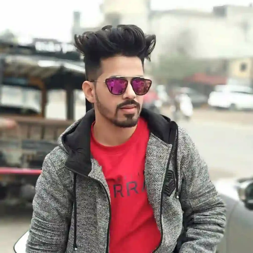 A young man with stylish hair wears shades and a red shirt under a gray jacket, posing outdoors with a blurred background.