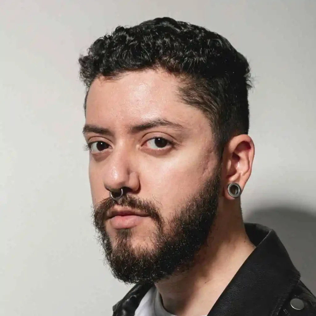 A person with medium-length curly black hair and a beard gazes intently at the camera, wearing a leather jacket.