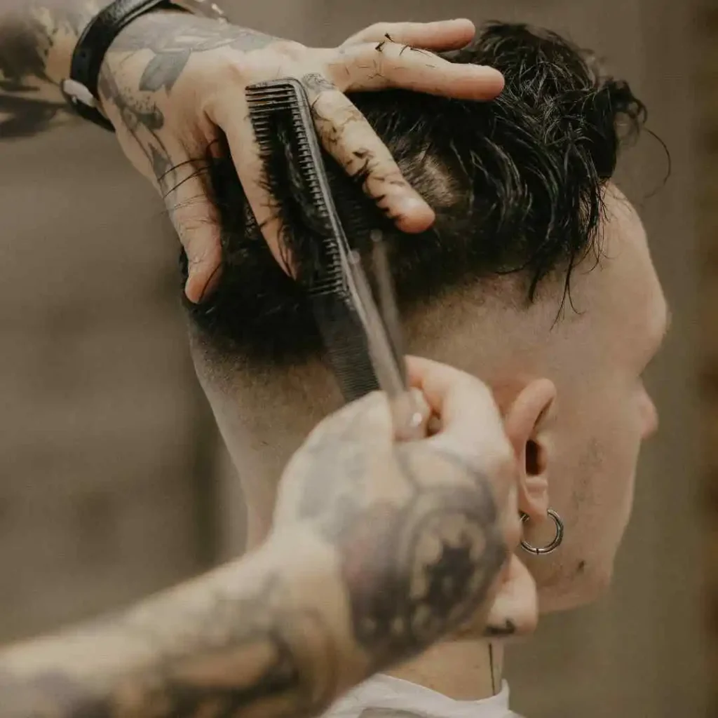 A barber styles a client's hair, using a comb and scissors, showcasing intricate tattoos on the barber's hands.