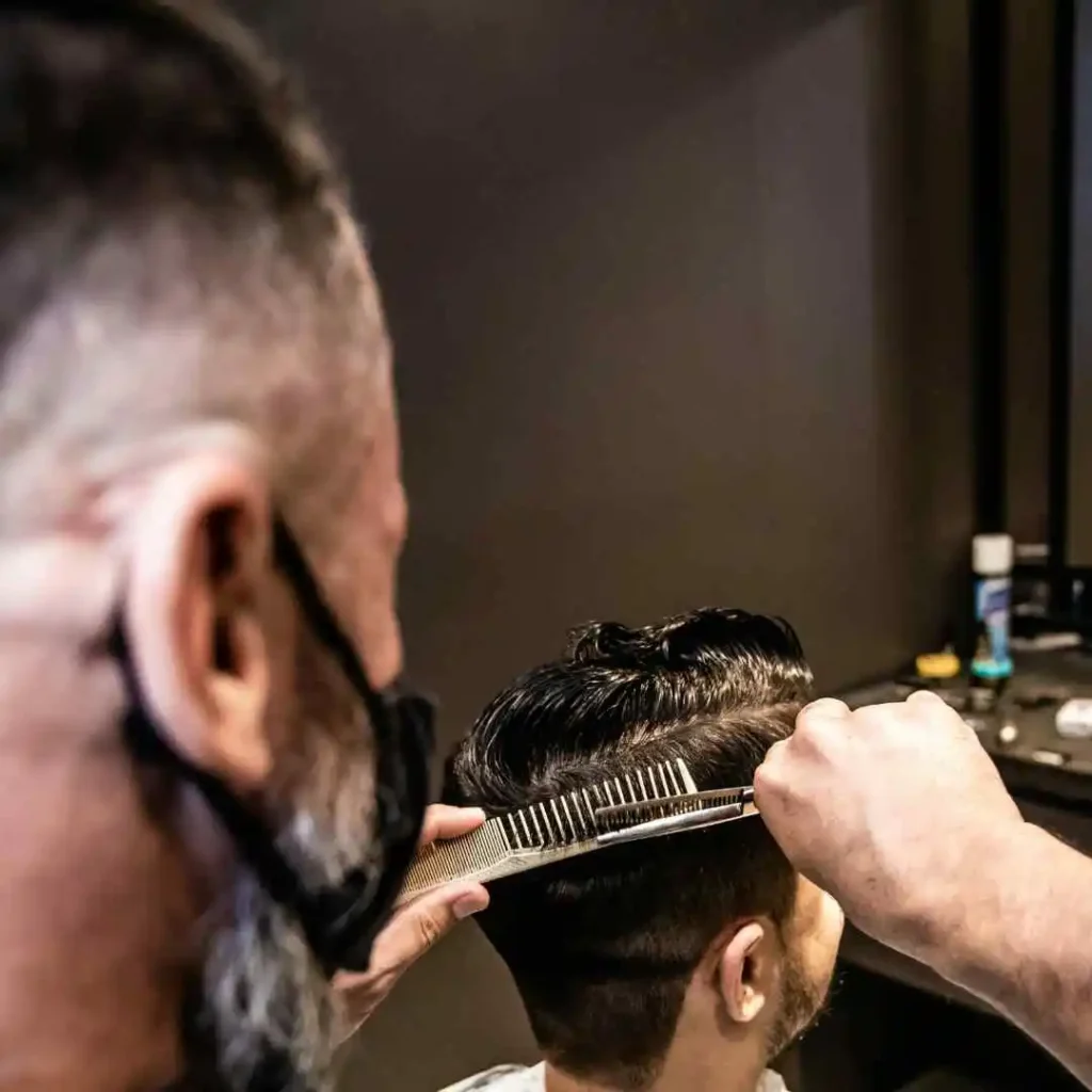 A barber, wearing a mask, styles a client's hair with a comb in a modern salon setting, focusing intently on their work.