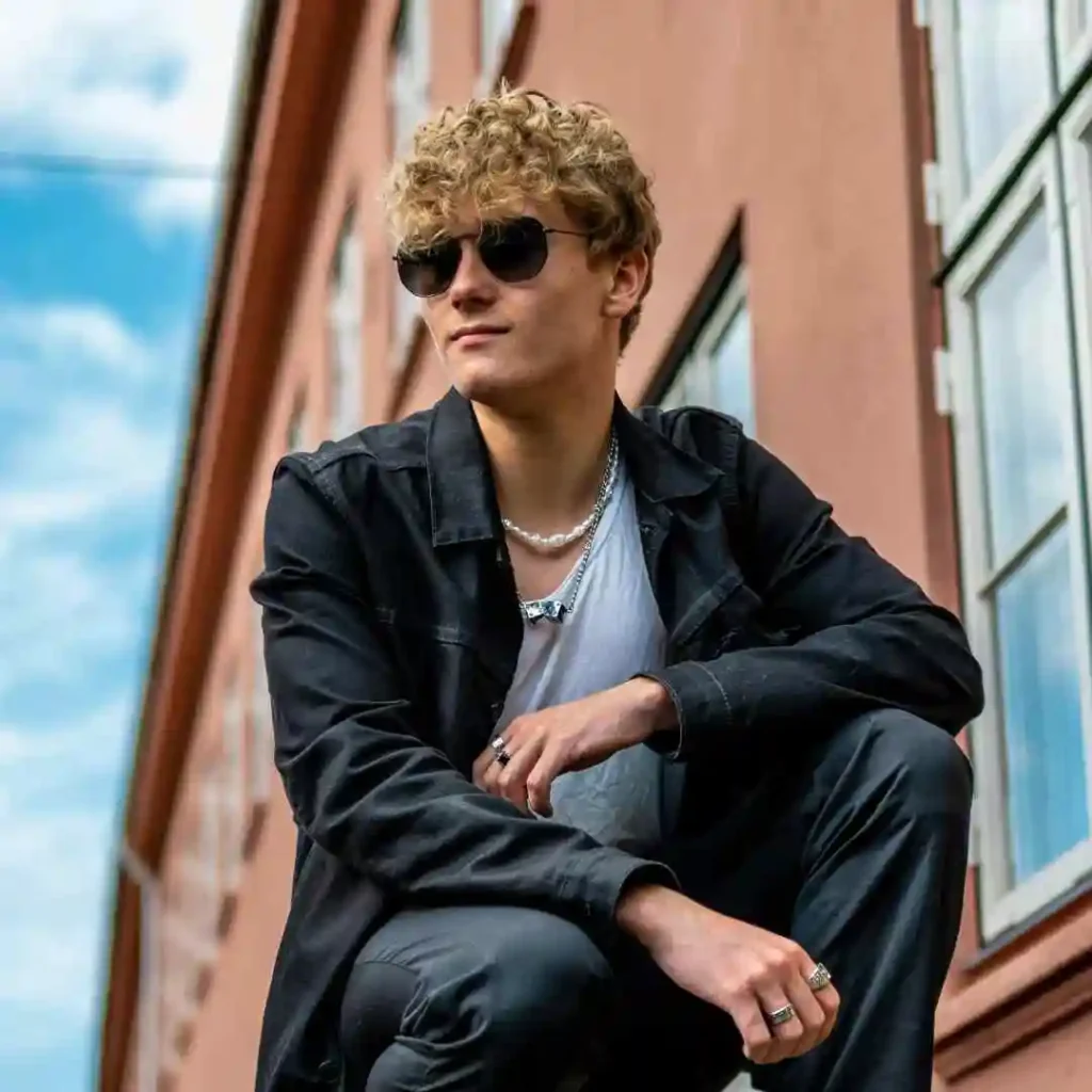 A man sitting on a ledge in front of a building, sporting a trendy men's short curly haircut.