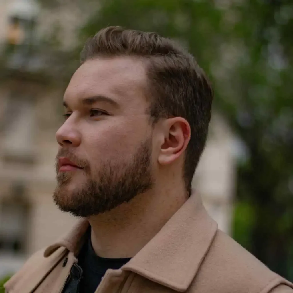 Man in a beige coat outdoors with a blurred background, sporting a quiff hairstyle.