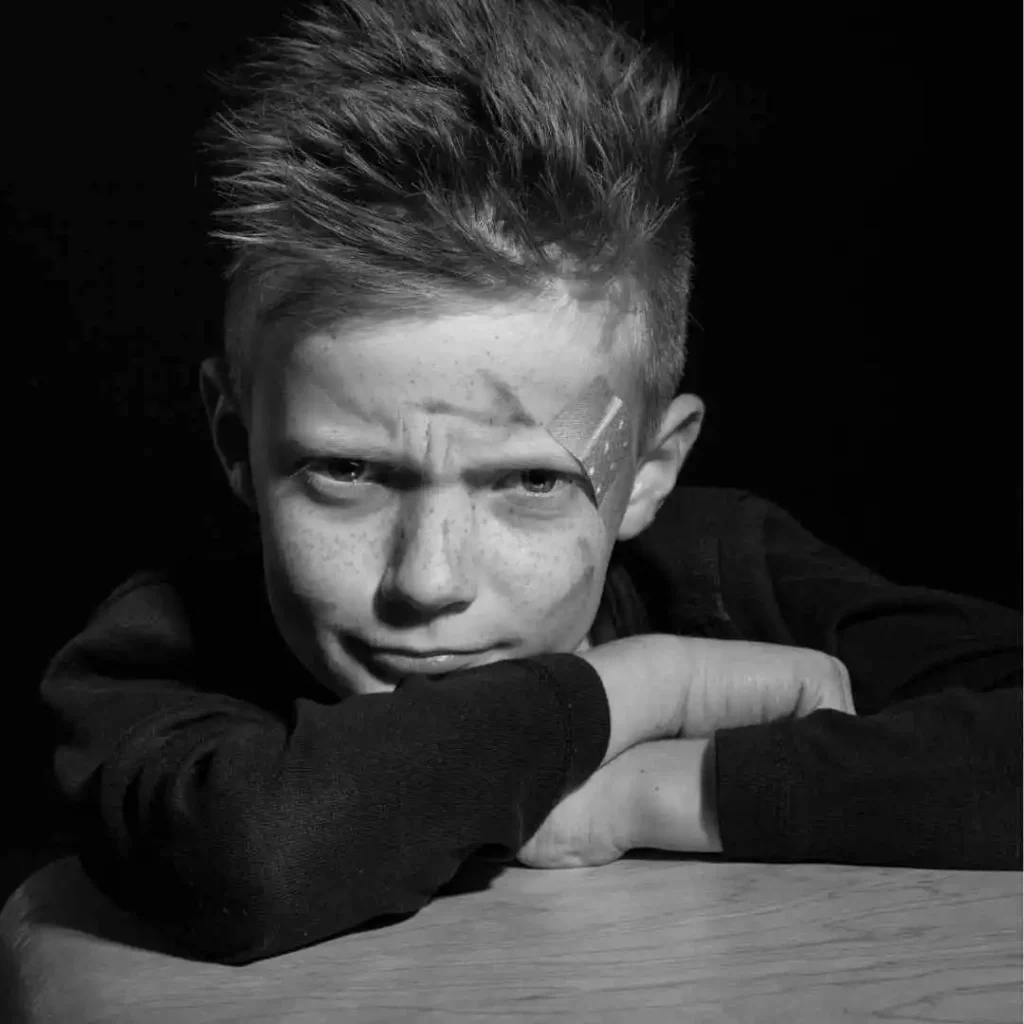 Man with a quiff hairstyle and spiky hair, leaning on a table with arms crossed, in monochrome.