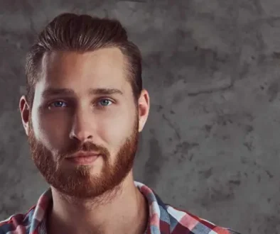 A young man with a full beard and blue eyes poses against a textured gray background, wearing a red and blue checkered shirt, showcasing a stylish Men's Crop & Crew Cut.