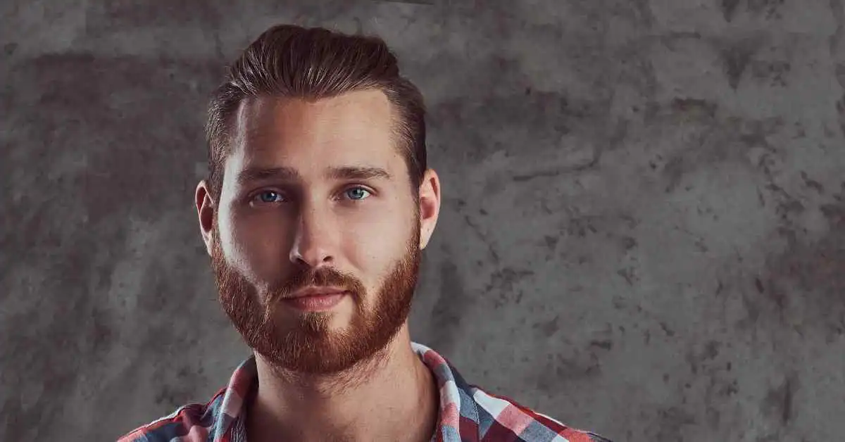 A young man with a full beard and blue eyes poses against a textured gray background, wearing a red and blue checkered shirt, showcasing a stylish Men's Crop & Crew Cut.