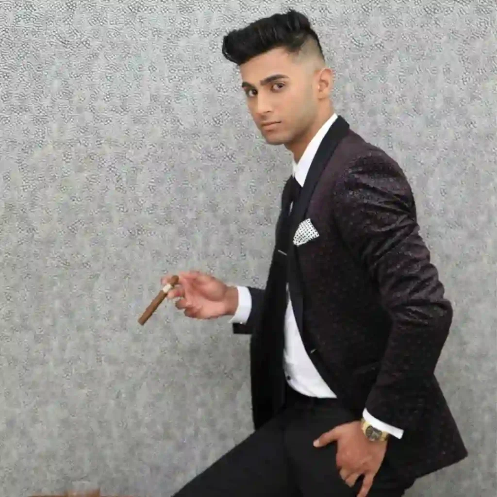 Man in a stylish black suit holding a cigar, standing against a textured wall, with a quiff hairstyle.