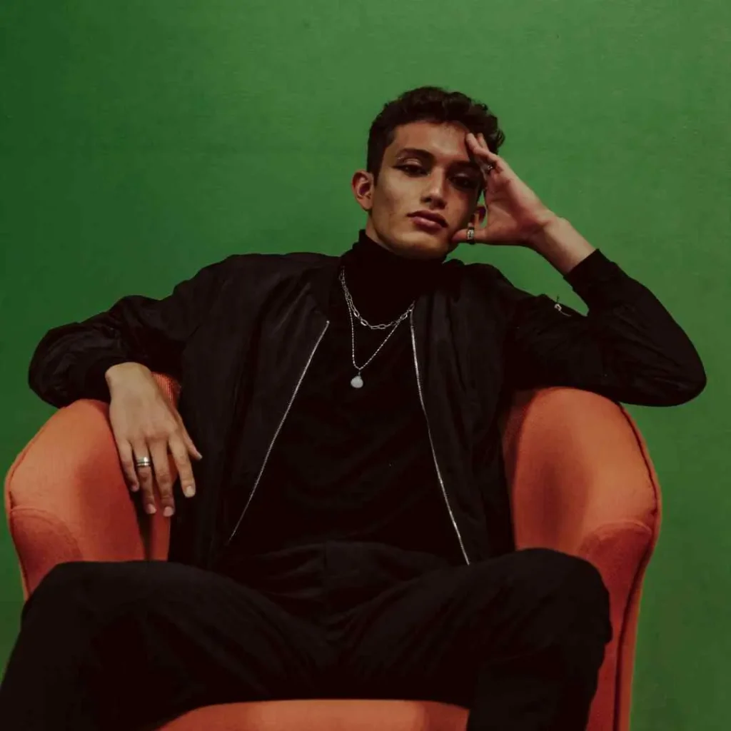 A man sitting in a chair with his hand on his head, featuring a stylish men's short curly haircut.