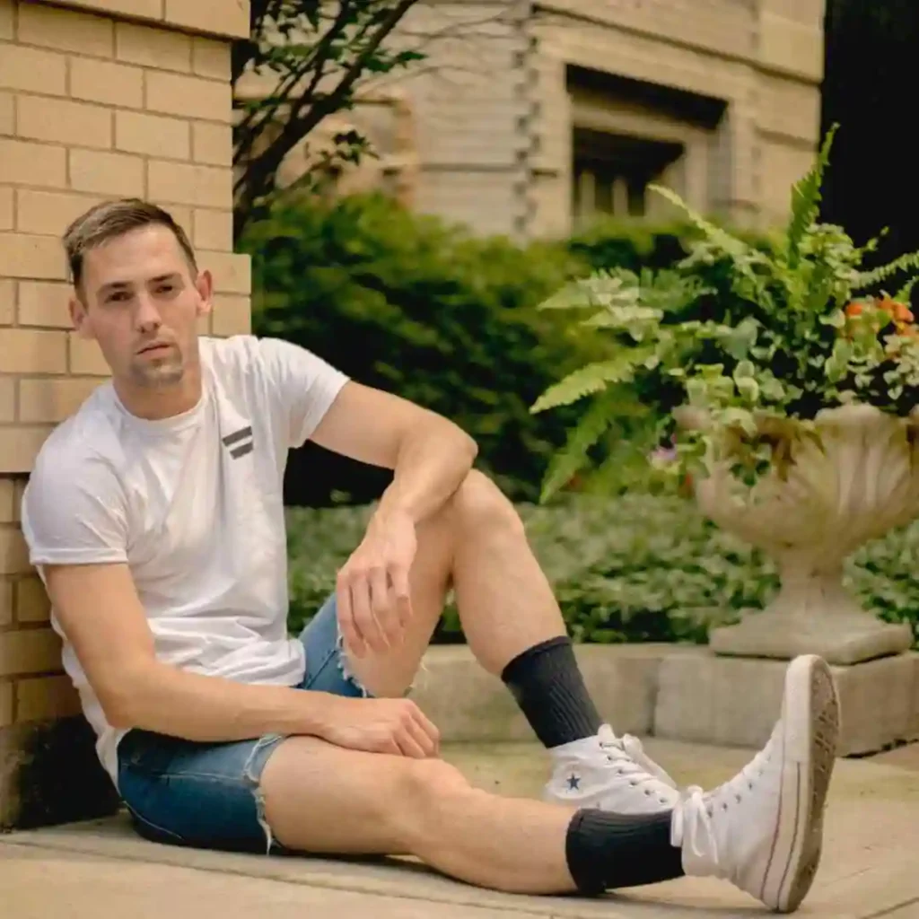 Man sitting on pavement by a building corner with shorts and sneakers, sporting a men’s hairstyle undercut.