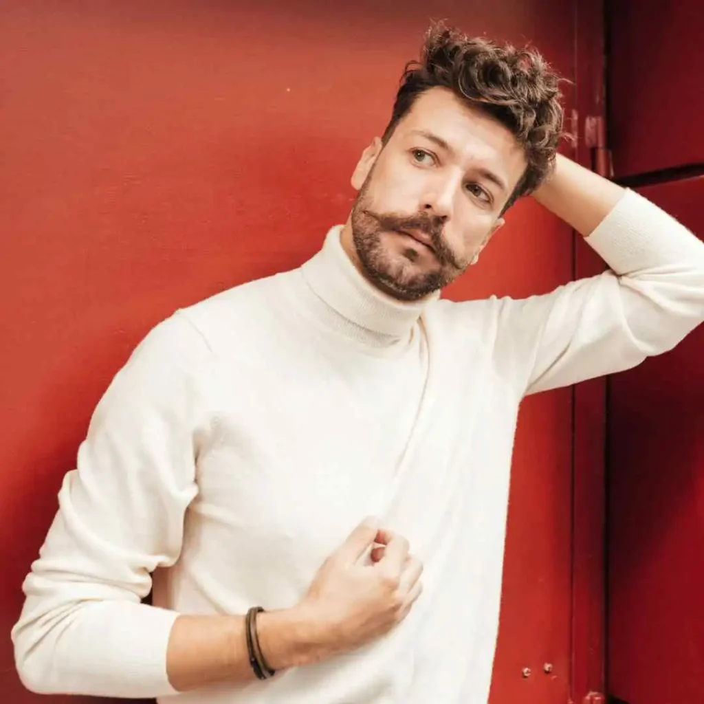 A person in a white sweater posing against a red background, sporting a quiff hairstyle.