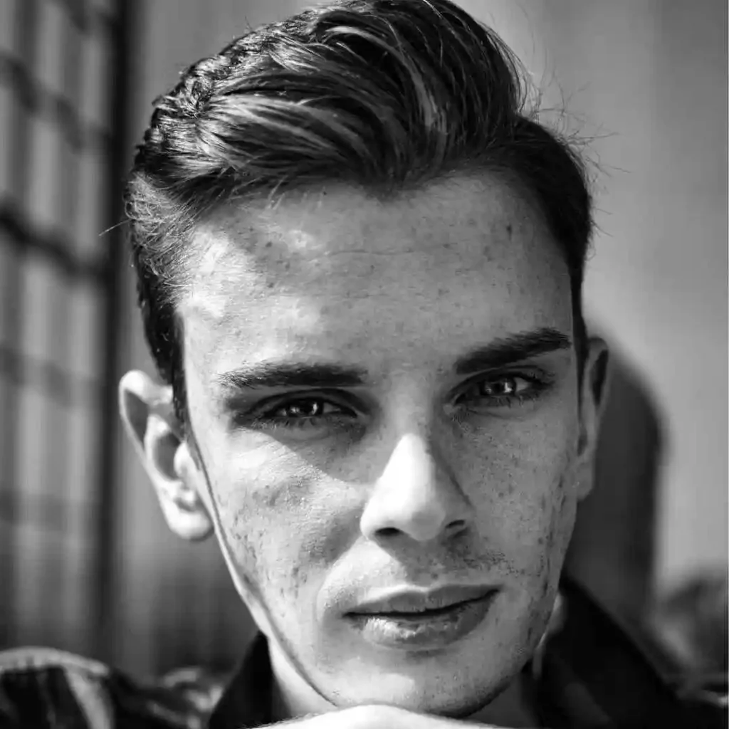 A black and white photo of a young man, featuring a stylish men's short curly haircut.
