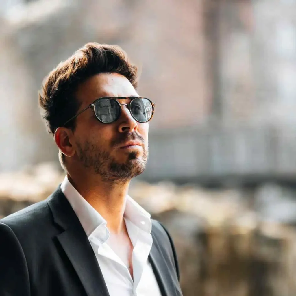 A man in a suit with spectacles, looking away to the right, showcasing a quiff hairstyle, with a blurry background.