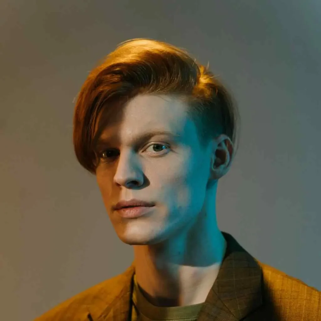 Person with slicked-back hair wearing a brown jacket and green shirt, with a gray background, featuring a quiff hairstyle.