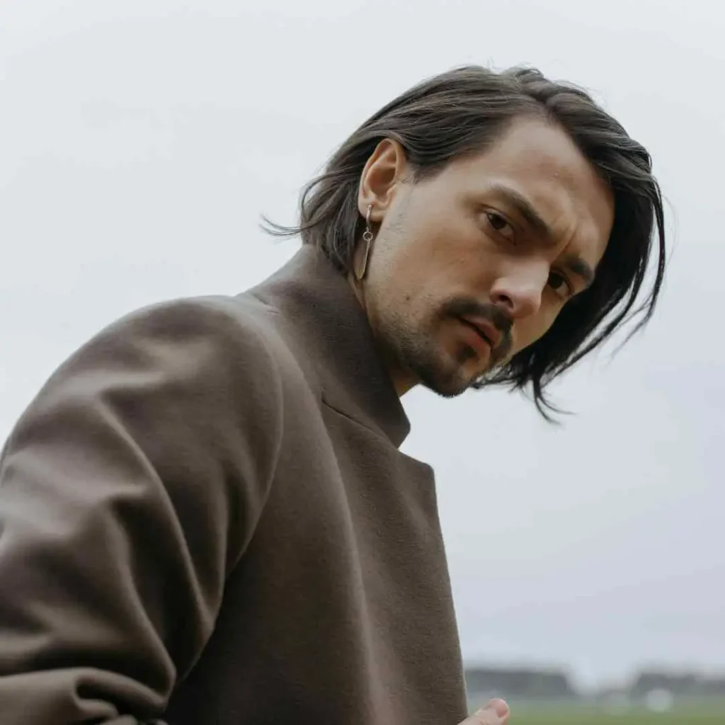 A stylish person in a brown coat with a visible earring, looking to the side, outdoors on a breezy day, sporting a men’s hairstyle undercut.