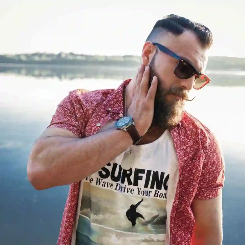 A man in a surfing t-shirt and red shirt by the water, with his face obscured, sporting stylish summer hairstyles for men.