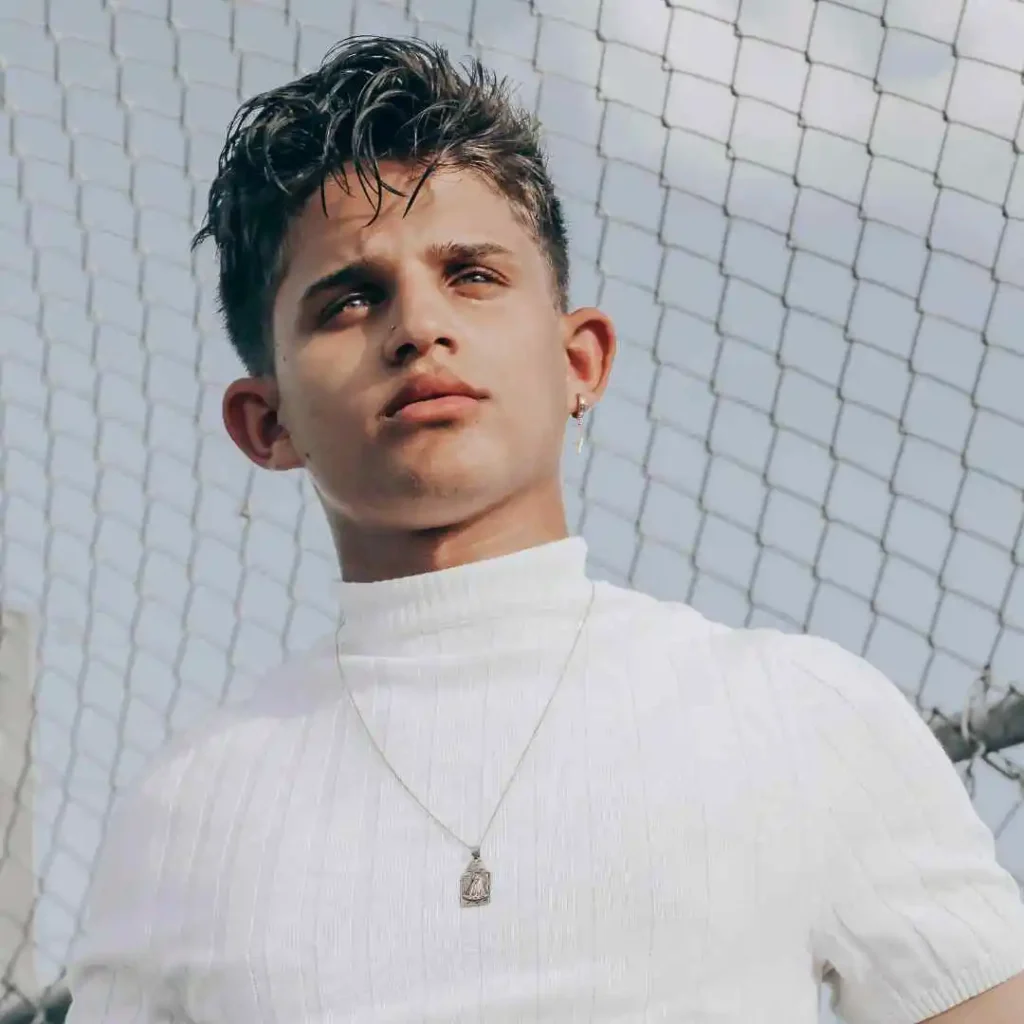 Person in white turtleneck and pendant with short hair against a chain-link fence.