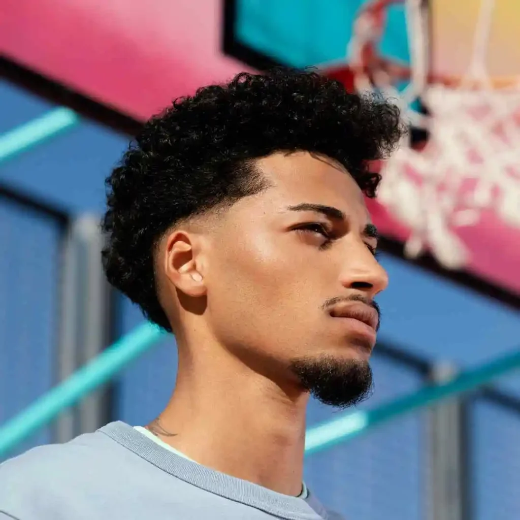 A close-up of a young man with fade hairstyles for men, standing in front of a basketball hoop in the background.