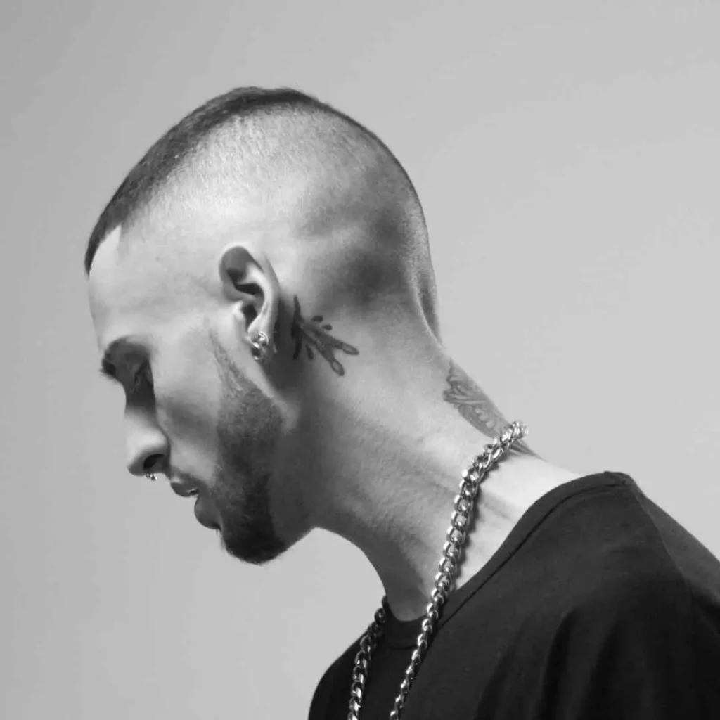 Profile of a person with a shaved head, neck tattoos, and a chain necklace. Black and white photo.