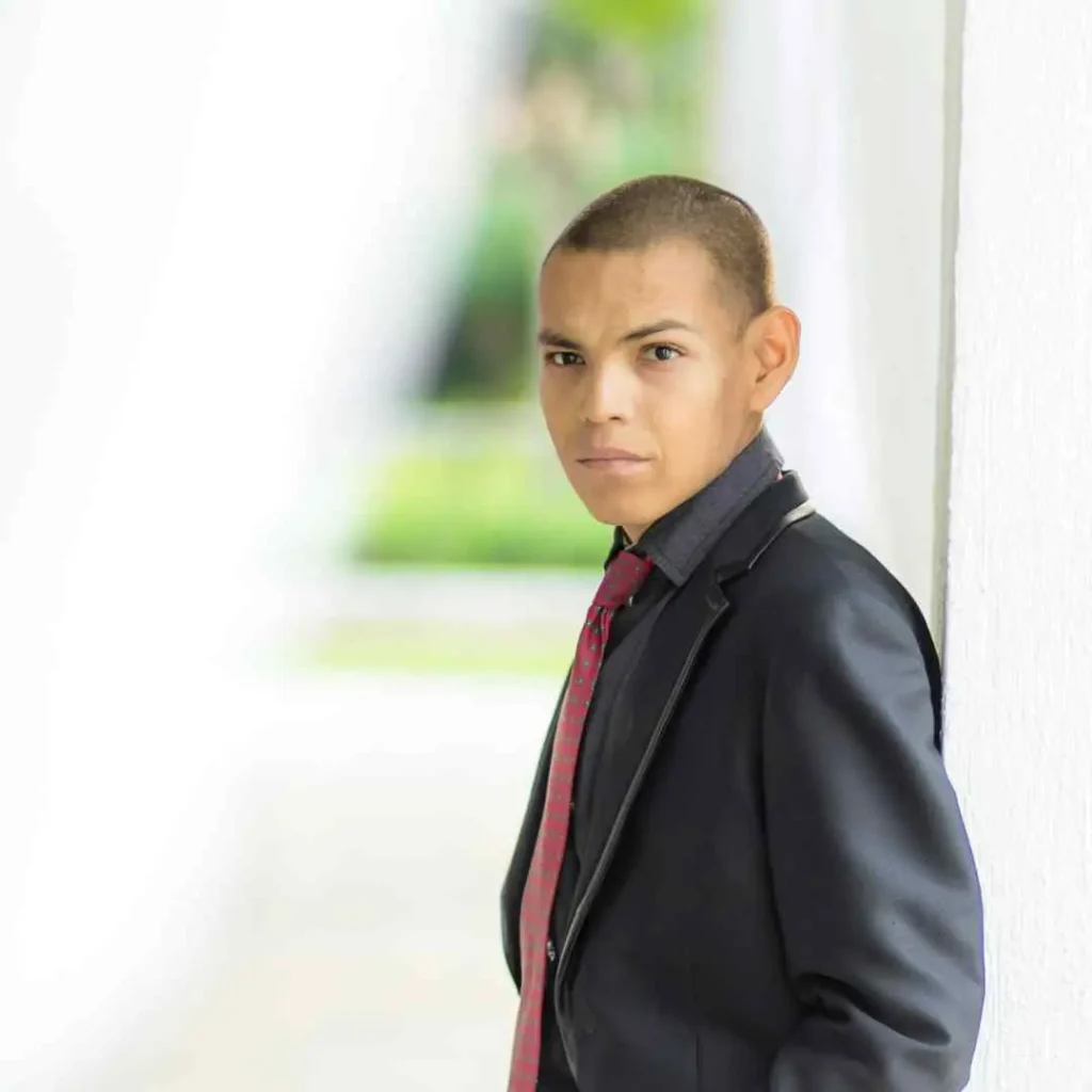 Person in suit and tie standing by a white wall with a greenery background. Teenage guy Haircuts style.