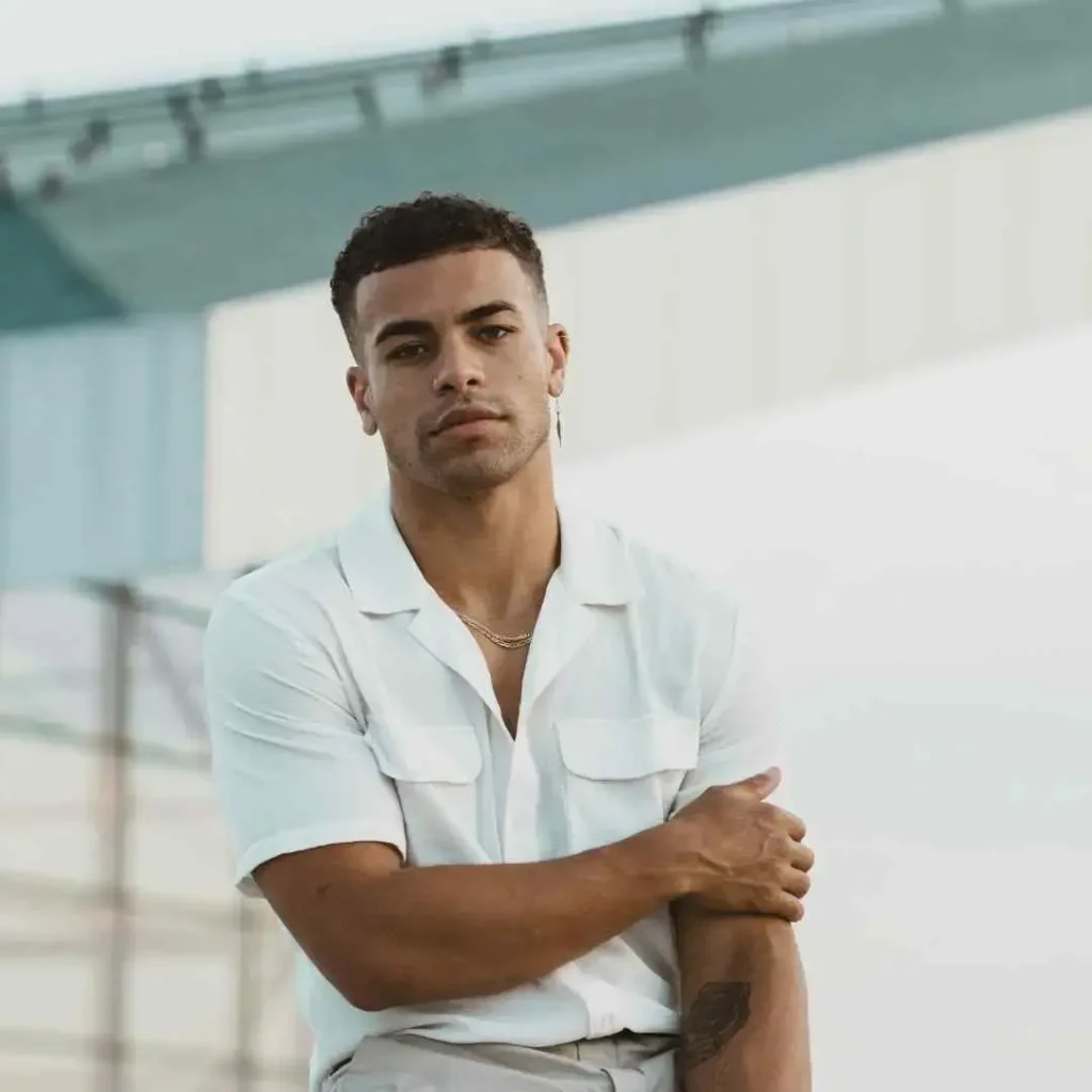 Man in a white shirt with crossed arms and a visible tattoo, sporting a men hairstyles buzz cut.