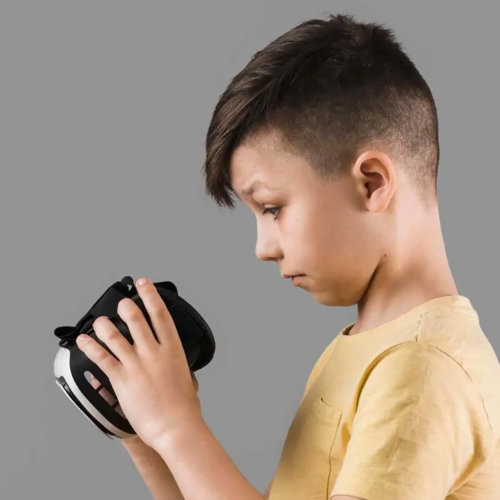 Youth in yellow shirt holding a black virtual reality headset against a grey background.