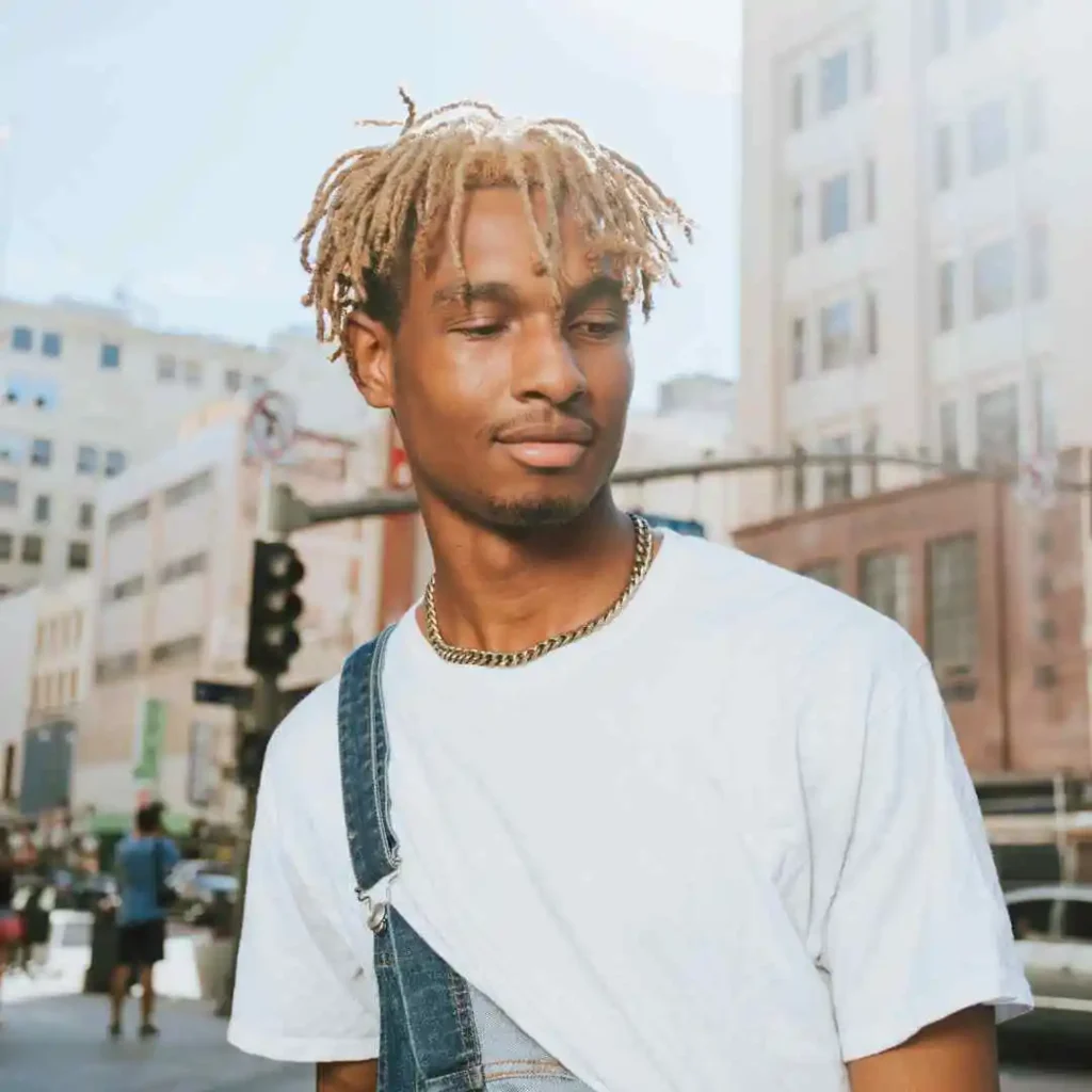 Person in a white tee and denim overalls against an urban street backdrop.