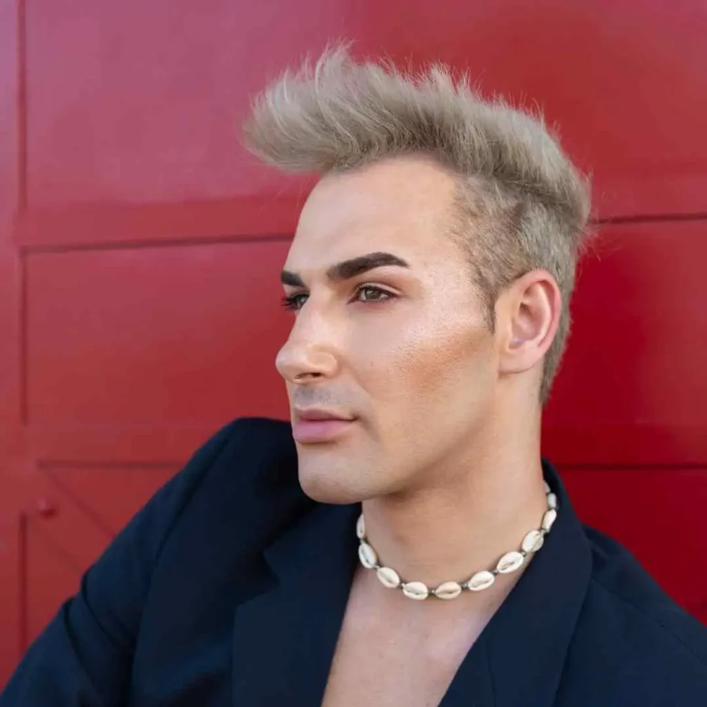 Person with blond hair in a black jacket and white necklace, with a red background.