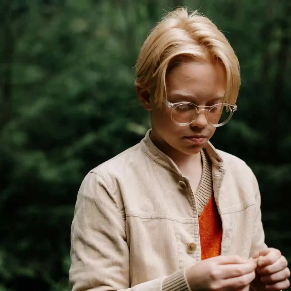 Person in a beige jacket over an orange shirt, standing before a greenery background.