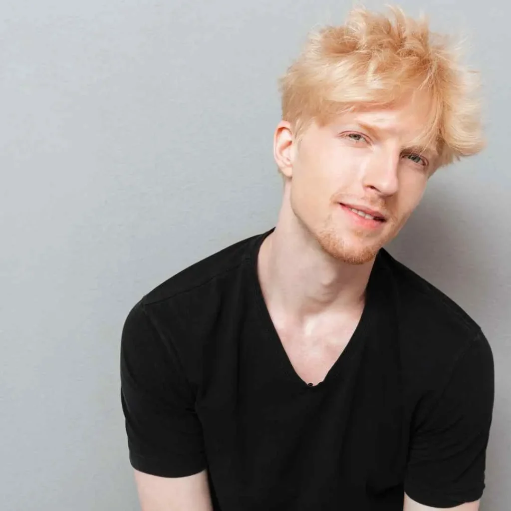 A man with blonde hair, wearing a black shirt, and sporting a pompadour hairstyle.