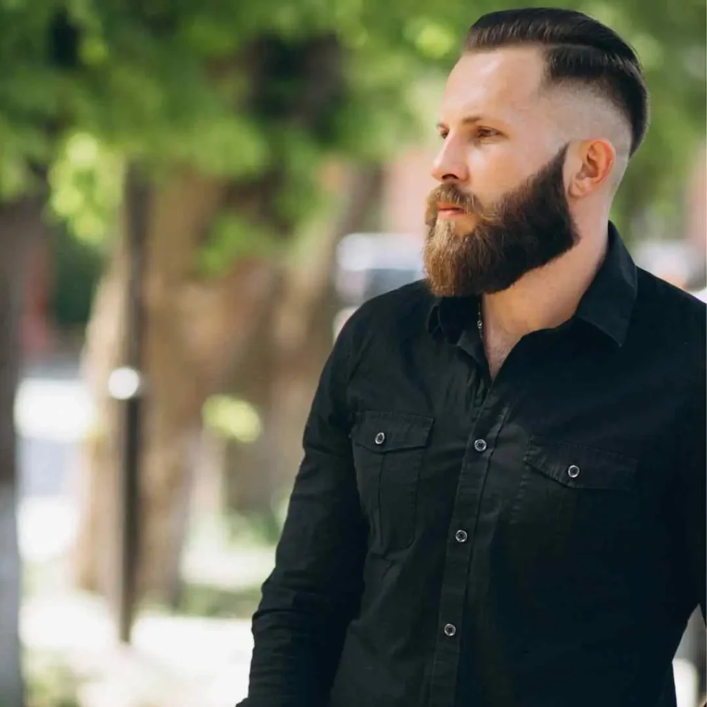 Man in a black shirt with a stylish undercut and beard, outdoors with greenery in the background, featuring a modern Mohawk men hairstyle.