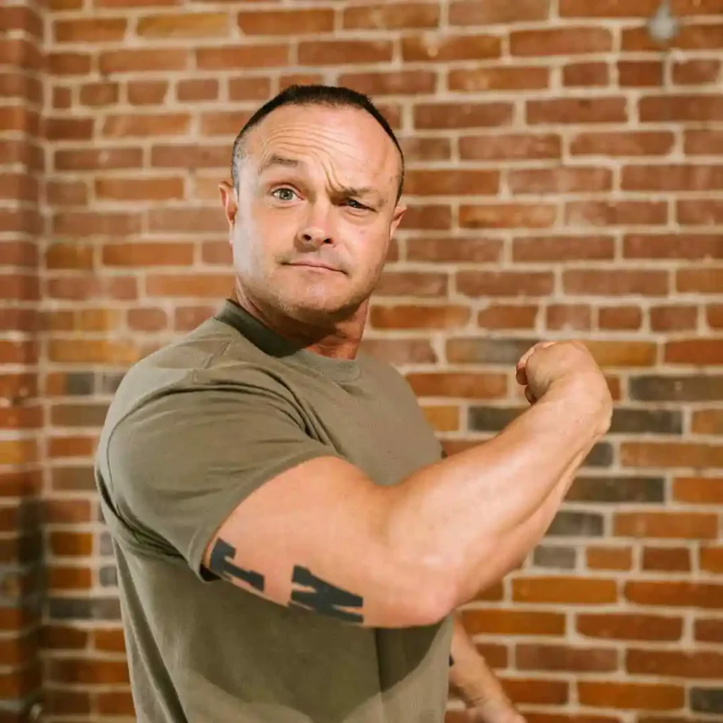 Man flexing his bicep in front of a brick wall, wearing a green t-shirt with a tattoo on his arm, showing off a men hairstyles buzz cut.