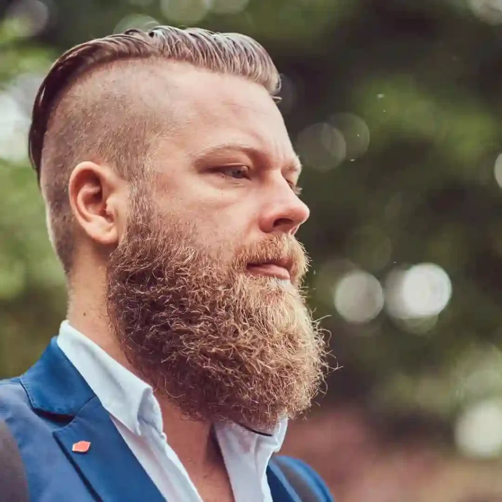 Man with a slicked-back hairstyle and a full beard, wearing a blue suit, exuding a polished and sophisticated look with a bold Mohawk men hairstyle.