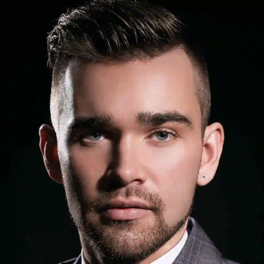 A side profile of a person with a modern hairstyle, wearing a grey blazer, against a black background.