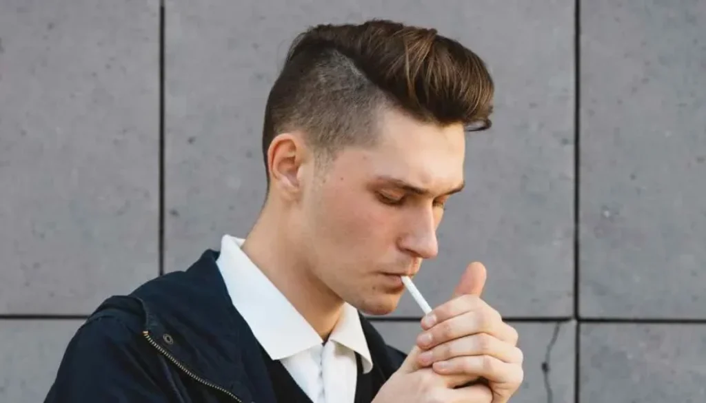 A man with a pompadour hairstyle smoking a cigarette in front of a wall.