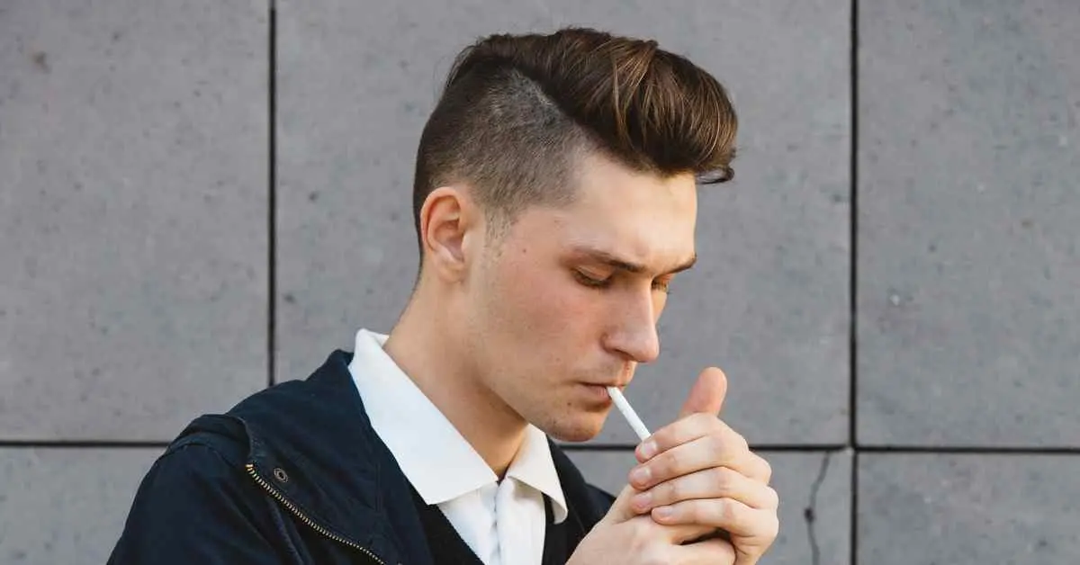 A man with a pompadour hairstyle smoking a cigarette in front of a wall.