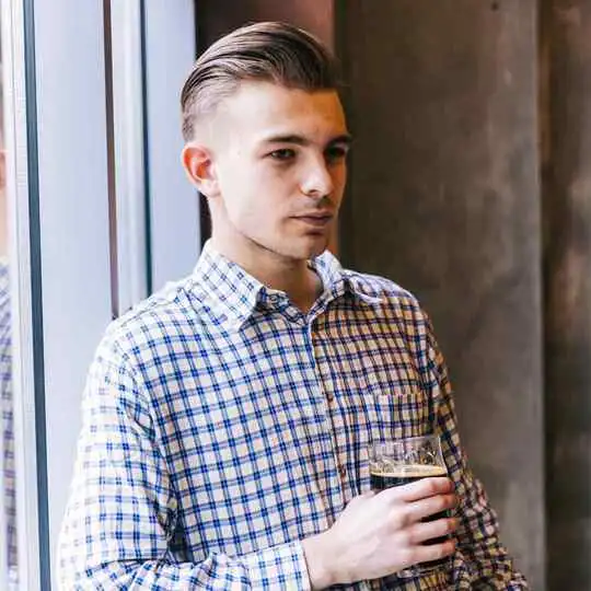 A man standing next to a window holding a drink and sporting a pompadour hairstyle.