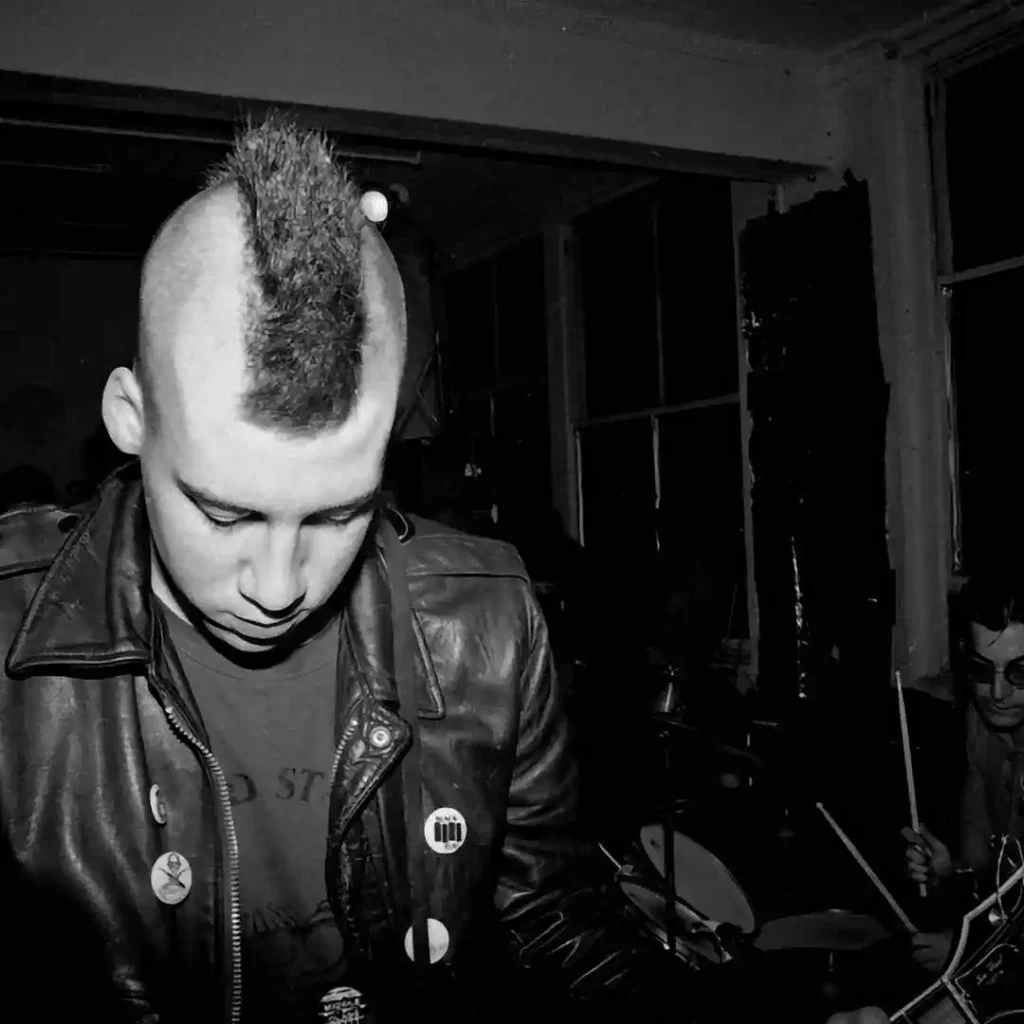 A person with a mohawk wearing a leather jacket adorned with buttons, in a dark room.