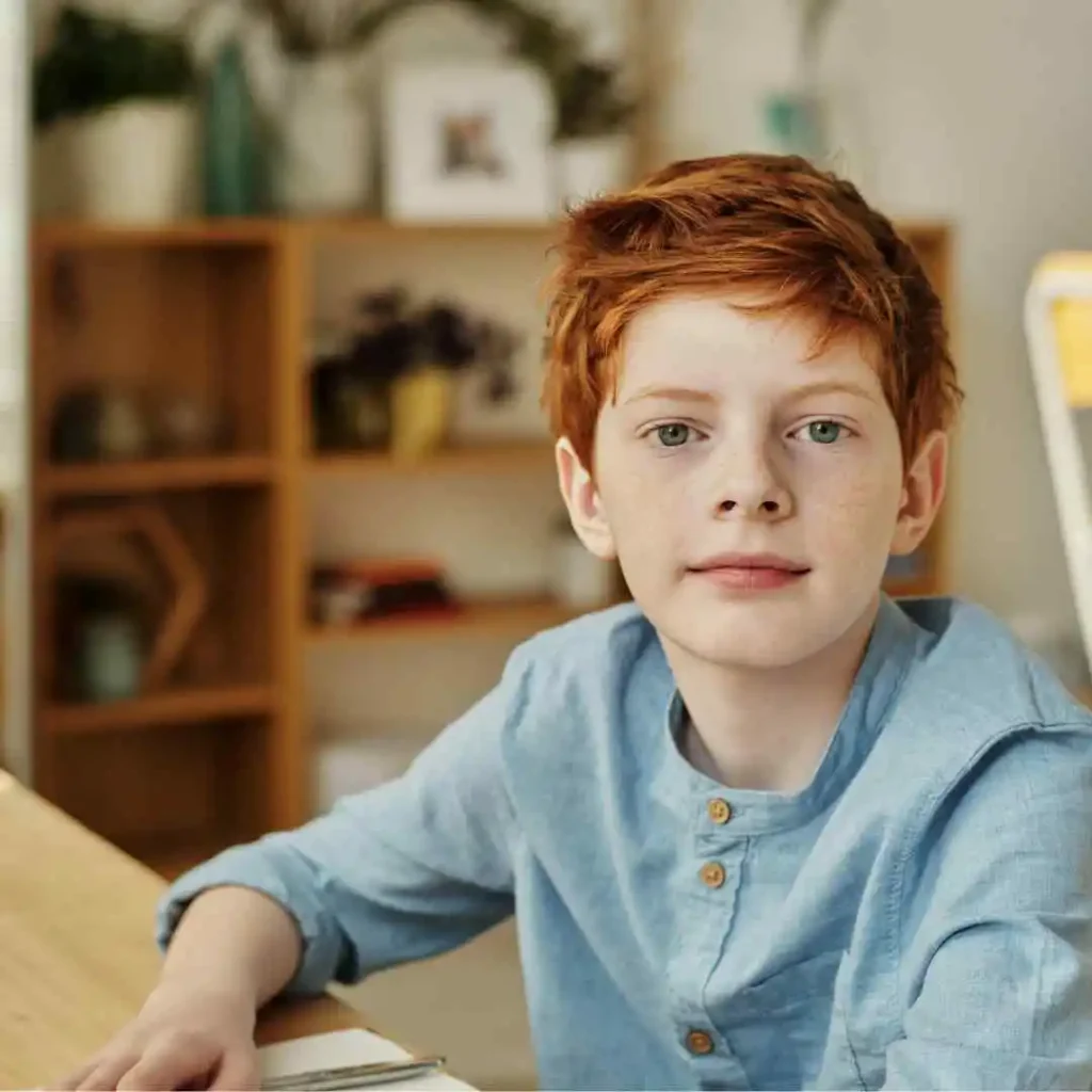 A child in a blue shirt sitting at a table with a book, in a room with a bookshelf.