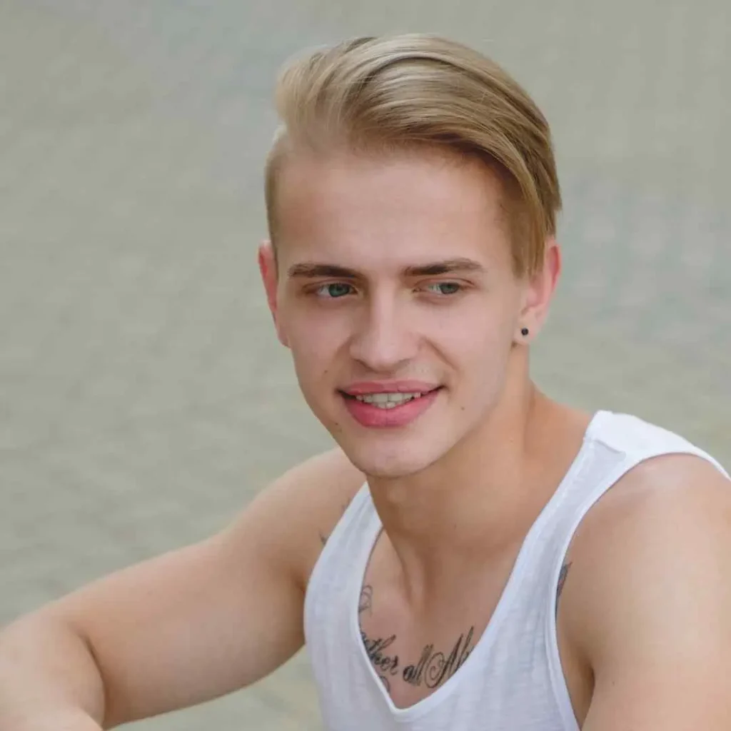 Person with a side-swept hairstyle and white tank top showing a tattoo on the collarbone.